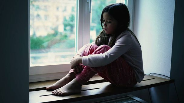 Melancholijna, smutna dziewczynka siedząca przy oknie mieszkania patrząca na widok w cichej kontemplacji. Przemyślane dziecko w głębokiej introspekcji umysłowej, przedstawiające samotność w dzieciństwie - Zdjęcie, obraz