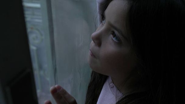 One pensive little girl leaning on glass window in deep mental introspection. Thoughtful childhood contemplation, child struggling with solitude at home - Photo, Image