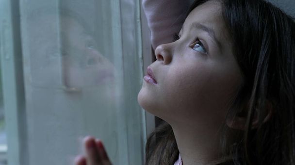 Sad little girl leaning on glass window with pensive expression, melancholic emotion of child struggling with loneliness and solitude - Photo, Image