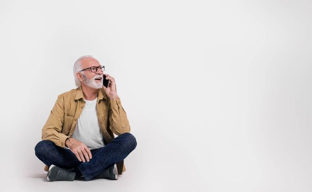 Oudere mannelijke professional praten over smartphone en weg te kijken tijdens het zitten op een witte achtergrond - Foto, afbeelding