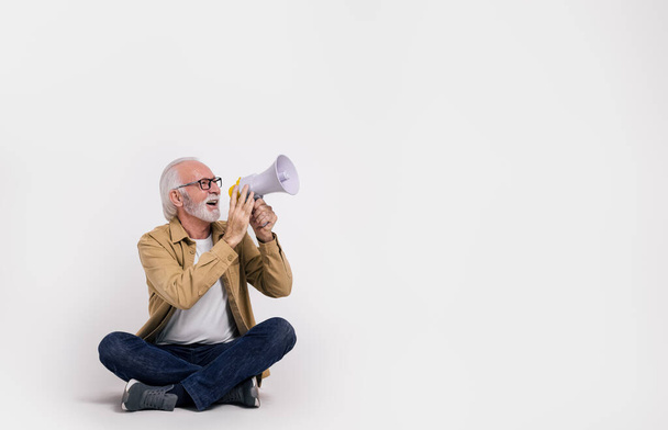 Старший мужчина-активист кричит над мегафоном и смотрит в сторону, сидя на белом фоне - Фото, изображение