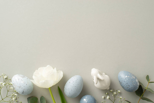 Tema de celebração da primavera na foto vista superior com ovos de ardósia acinzentada, uma estatueta de coelho, gypsophila, tulipa e eucalipto dispostos em uma tela cinza macia, com um vazio para texto ou publicidade - Foto, Imagem