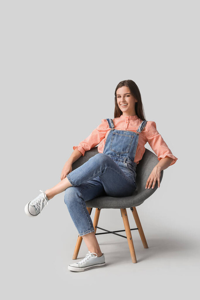 Krásná šťastná mladá žena sedí na moderní židli proti šedému pozadí - Fotografie, Obrázek