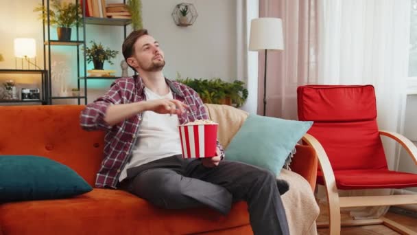 Emocionado hombre comiendo palomitas de maíz viendo interesante juego de deportes en serie de televisión en línea contenido de la película de medios sociales sentado en el sofá en la habitación en casa. Feliz chico caucásico en camisa a cuadros disfrutando de la película durante el fin de semana - Imágenes, Vídeo