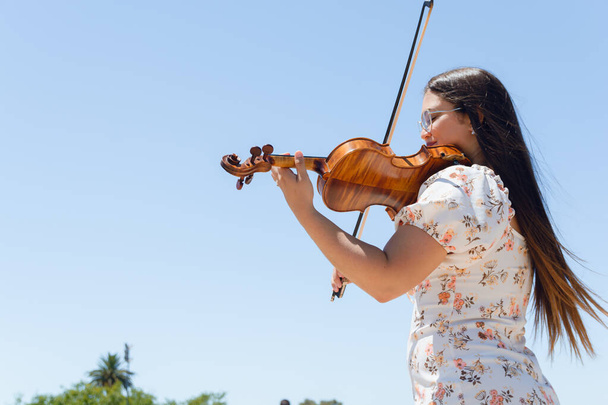 νεαρή busker latin γυναίκα με μακριά μαλλιά στο βιολιστικό φόρεμα κάνει παρουσίαση σε εξωτερικούς χώρους παίζοντας βιολί, με τον ουρανό στο παρασκήνιο και να αντιγράψετε χώρο. - Φωτογραφία, εικόνα