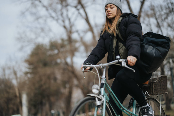 Mulher adulta ativa monta sua bicicleta em um parque em um dia frio, carregando uma bolsa grande, promovendo viagens ecológicas e fitness. - Foto, Imagem