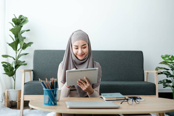 Junge Business Asian Business Muslimische Frau im Hidschab, die mit einem Tablet am Fenster im Wohnzimmer sitzt. Hochwertiges Foto - Foto, Bild