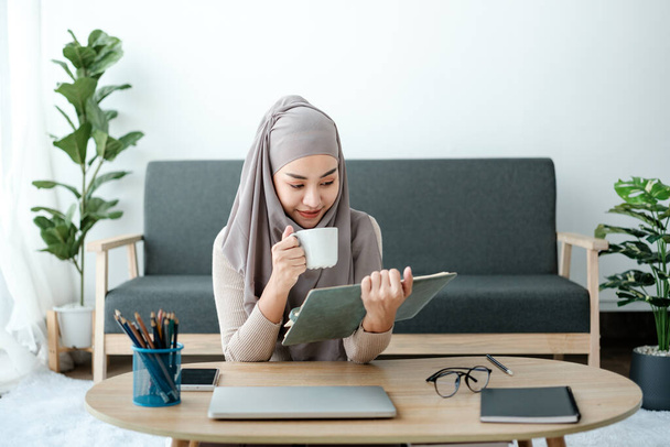 Eine attraktive und glückliche junge asiatisch-muslimische Frau genießt ihren Kaffee, während sie im Wohnzimmer ein Buch liest. Heimische Lebens- und Freizeitkonzepte. Hochwertiges Foto - Foto, Bild