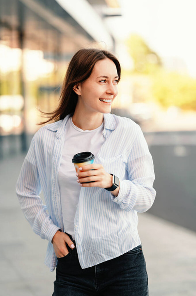 Vertikales Foto einer fröhlichen jungen Frau, die lässig durch die Straßen der Stadt läuft und Kaffee to go in der Hand hält. - Foto, Bild