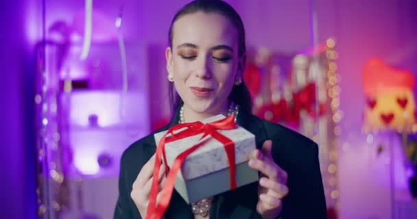 Feliz joven mujer sosteniendo envuelto caja de regalo mientras está de pie en casa durante la celebración del día de Galentines - Imágenes, Vídeo