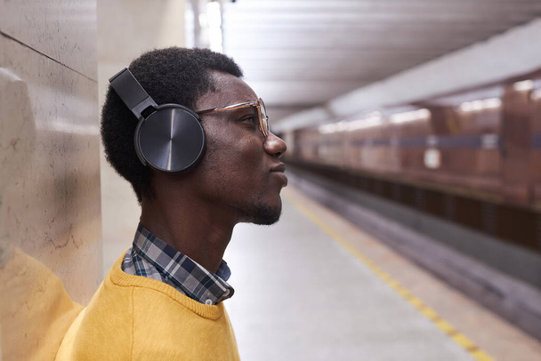 Vista lateral do homem em fones de ouvido sem fio ouvindo música em pé no subsolo - Foto, Imagem