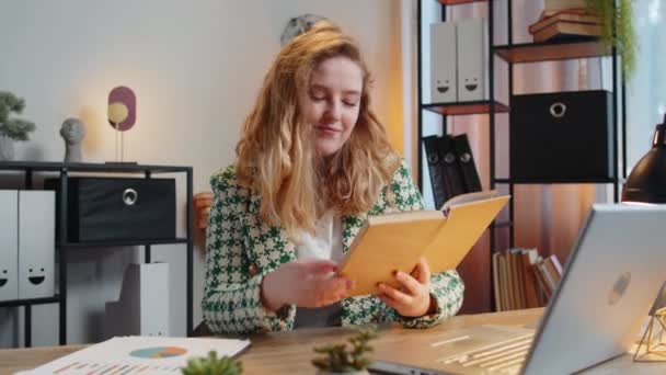 Kaukázusi fiatal üzletasszony érdekes könyvet olvas, lapozgat mosolyogva élvezi az irodalmat, munka után pihenőt tart. Boldog vidám szabadúszó lány laptop pihentető otthoni irodai asztal. - Felvétel, videó
