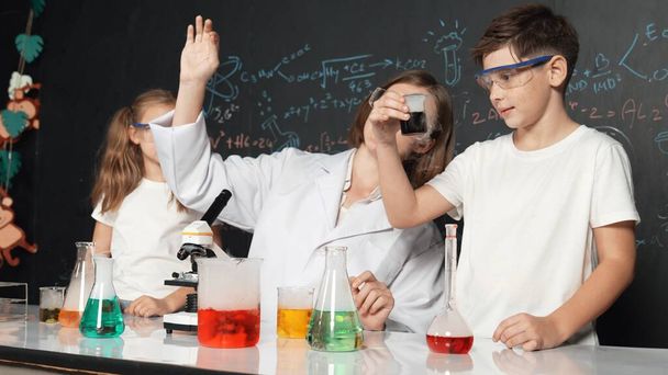 Кавказький хлопчик змішує хімічну рідину, коли вчитель дає поради. Професійний інструктор у лаборантському костюмі шукає різноманітного студента за столом зі склянкою, наповненою кольоровим рішенням. Руйнування. - Фото, зображення