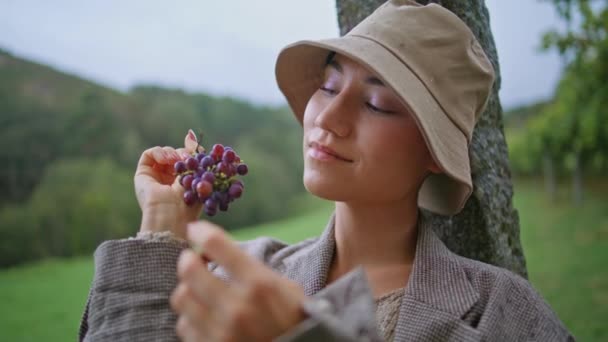 Szekrényes nő szőlőt eszik a borús őszi szőlőskertben. Portré gyönyörű lány csukott szemmel élvezi a friss bogyók ízét ültetvényen. Trendi hölgy Panamában ízletes lédús szőlő szüret. - Felvétel, videó