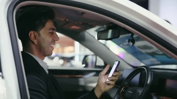 Pomyślny biznesmen siedzi w swoim własnym samochodzie i przewijania, oglądając śmieszne filmy na smartfonie. Szczęśliwy młody właściciel samochodu siedzi w samochodzie i jest szczęśliwy z zakupu, Korzystanie z telefonu komórkowego - Materiał filmowy, wideo