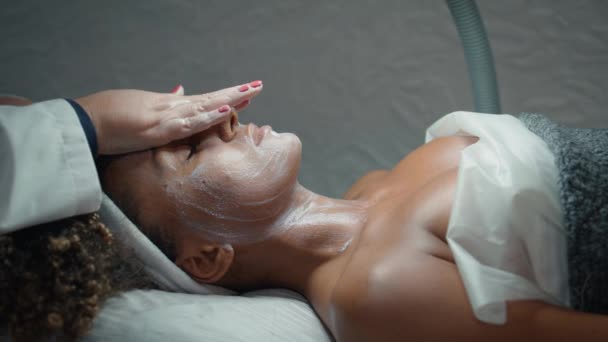 Καλλυντικά χέρια μασάζ προσώπου με θρεπτική κρέμα σε spa σαλόνι closeup. Χαλαρός Αφροαμερικάνος που απολαμβάνει τη διαδικασία κοσμετολογίας. Άγνωστος αισθητικός θεραπεία του δέρματος μετά την απολέπιση αναζωογόνησης. - Πλάνα, βίντεο