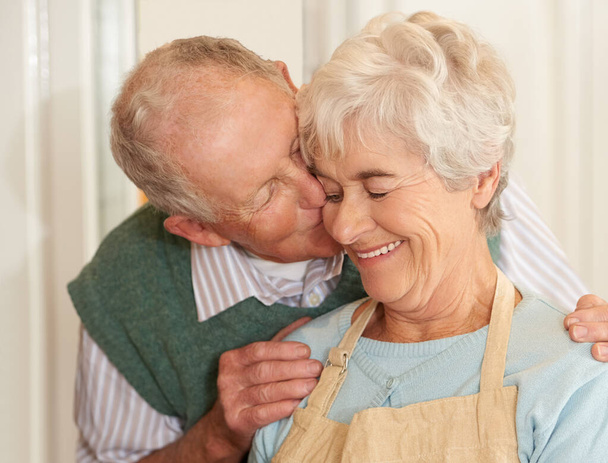 Старша пара, романтика і поцілунок вдома, посмішка і зв'язок на пенсії. Літні люди, люблять і щасливі у шлюбі або стосунках, у вихідні та підтримці прихильності або догляду на кухні. - Фото, зображення