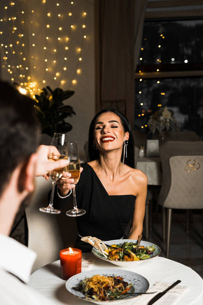 Ylellinen nainen mustassa mekossa on illallinen ravintolassa, jossa on kukkia ja lasi samppanjaa.Romanttinen illallinen. - Valokuva, kuva