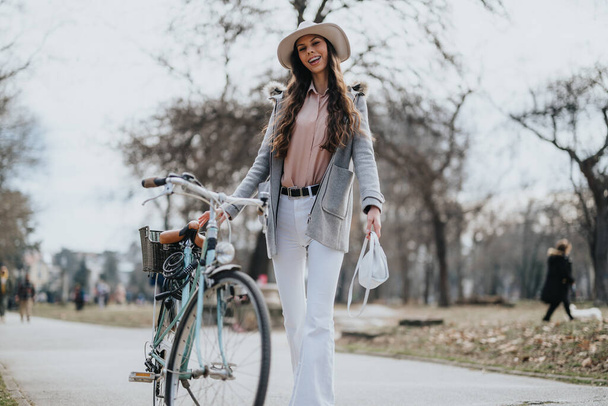 Μια κομψή νεαρή γυναίκα με καπέλο περπατάει με το παλιό της ποδήλατο σε ένα πάρκο, αποπνέοντας επιτήδευση και ευτυχία την άνοιξη.. - Φωτογραφία, εικόνα