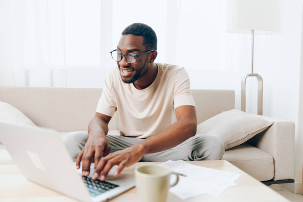 Улыбающийся афроамериканец-фрилансер, печатающий на ноутбуке, работающий из дома в современной гостиной Молодой черный парень сидит на уютном диване, окруженный учебниками и ноутбуком, полностью - Фото, изображение