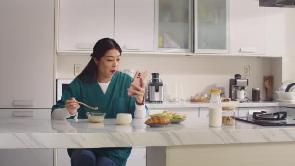 Mittlere Langzeitaufnahme einer fröhlichen Krankenschwester, die zu Hause frühstückt und Videos auf dem Smartphone anschaut - Filmmaterial, Video