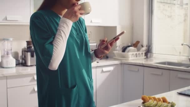 Döntse fel lövés a fiatal felnőtt nővér orvosi egyenruhában ivás tea és okostelefon otthoni konyhában - Felvétel, videó