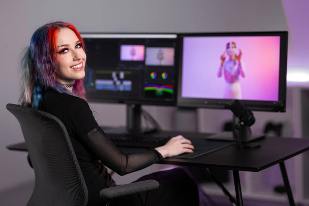 Mosolygó női professzionális kreatív digitális művész elmerül a vágás videó nő VR szemüveg, körülvéve élénk fények kortárs irodai térben. - Fotó, kép