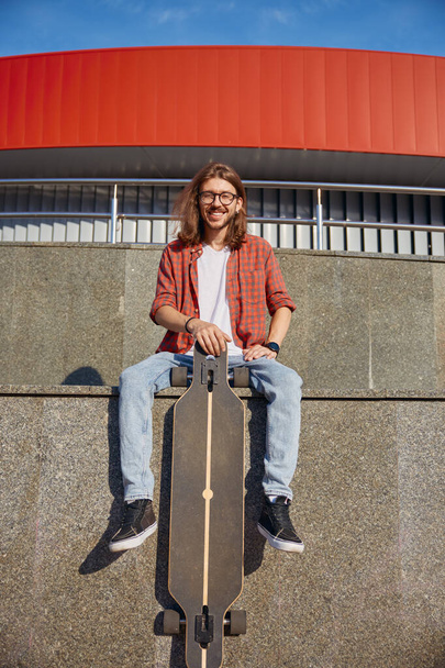 Porträt eines stilvoll lächelnden männlichen Hipster-Skateboarders, der im städtischen Stadtpark gut trainiert. Schöner Mann sitzt auf Brüstung mit Longboard - Foto, Bild