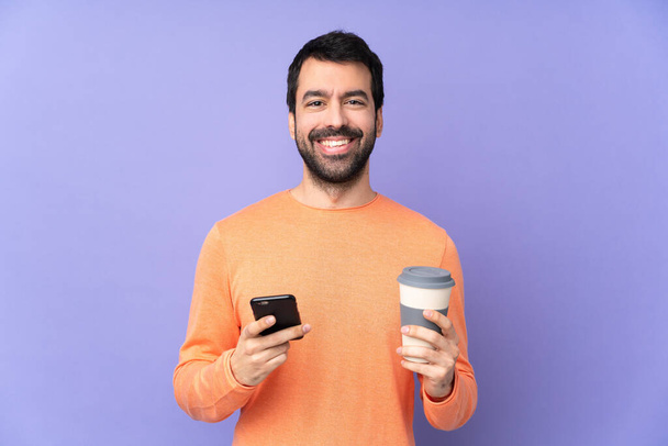 Caucásico guapo hombre sobre aislado púrpura fondo celebración de café para llevar y un móvil - Foto, Imagen