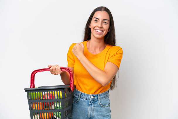 Young caucasian woman holding a shopping basket full of food isolated on white background celebrating a victory - Photo, image