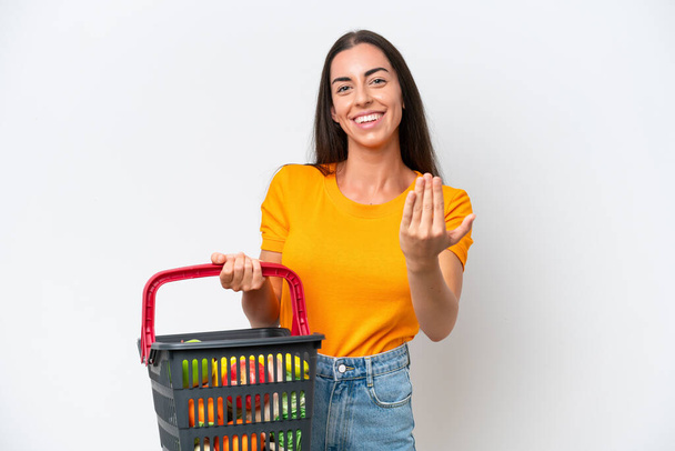 Young caucasian woman holding a shopping basket full of food isolated on white background inviting to come with hand. Happy that you came - 写真・画像