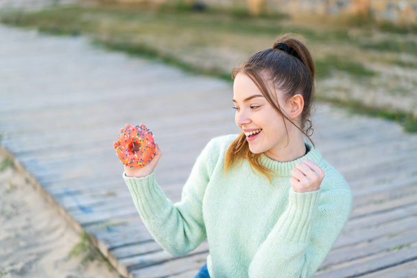 Ένα νεαρό όμορφο κορίτσι που κρατάει ένα ντόνατ στην ύπαιθρο γιορτάζοντας μια νίκη - Φωτογραφία, εικόνα
