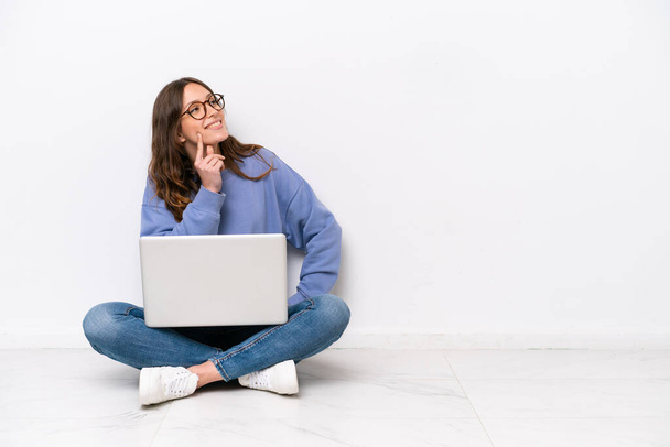 jonge Kaukasische vrouw met een laptop zitten op de vloer geïsoleerd op witte achtergrond denken een idee terwijl opkijken - Foto, afbeelding