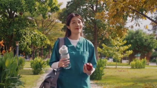 Mediana toma de joven trabajador médico alegre en uniforme sosteniendo agua y manzana yendo a la clínica en el parque de verano - Metraje, vídeo
