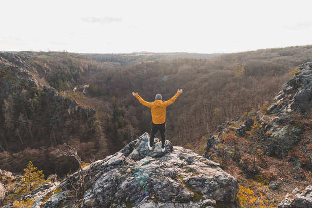 Podróżnik w żółtej kurtce stojący na skraju skały, cieszący się chwilą relaksu i widokiem na dolinę Divoke sarky, Praga, Czechy. Osiągnięcie sukcesu. - Zdjęcie, obraz