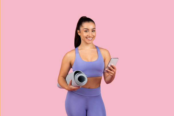 Счастливый любитель фитнеса в наборе для тренировок lilac держит коврик для йоги под одной рукой и смартфон в другой, готовый к тренировке, розовый рюкзак - Фото, изображение