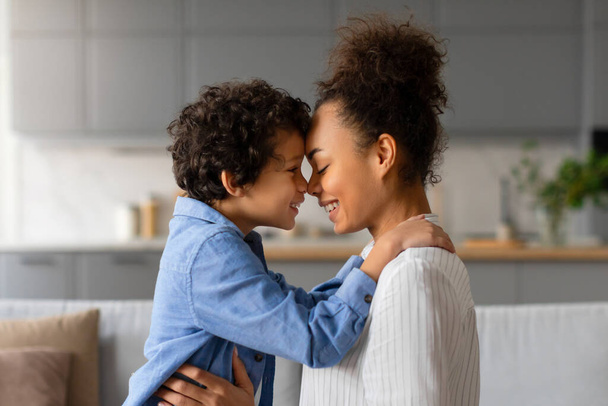 Affectueuse jeune mère noire avec les cheveux bouclés et le fils toucher doucement les fronts, exprimant l'amour et la connexion dans une atmosphère chaleureuse et accueillante, vue latérale - Photo, image
