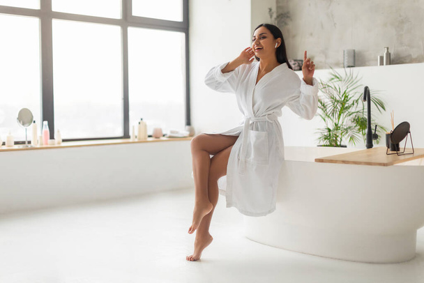 Indian woman in white bathrobe singing songs, wearing wireless earphones, cheerful eastern lady sitting on bathtub, relaxing in sunny bathroom interior, copy space, full length - Photo, Image