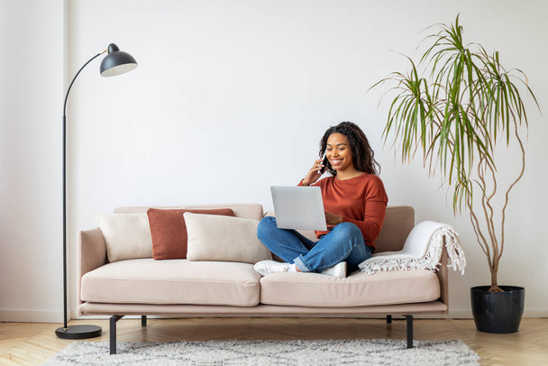 Koncept online práce. Mladá černošky žena pracující s notebookem a mluvení na mobilním telefonu doma, Afroameričanka pomocí počítače pro volné noze, sedí na gauči v obývacím pokoji, kopírovat prostor - Fotografie, Obrázek