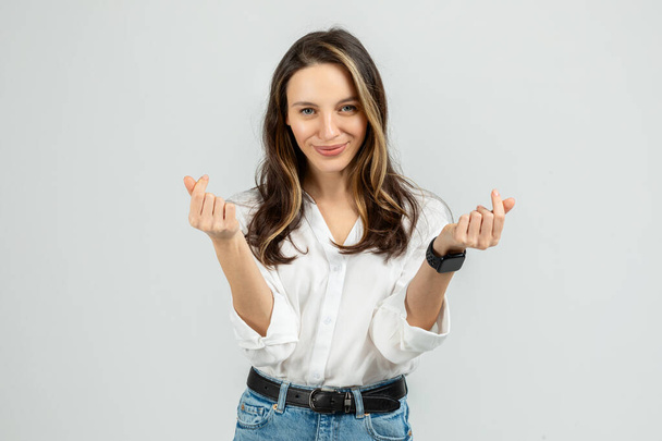 Брюнетка європейська жінка з щирою посмішкою робить грайливий грошовий жест з пальцями, одягнений у білу блузку і блакитні джинси, символізуючи фінансовий успіх або витрати - Фото, зображення