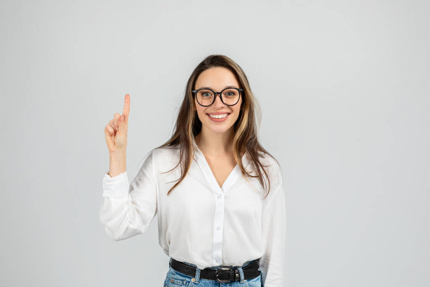 Σίγουρη χαρούμενη Ευρωπαία νεαρή γυναίκα με γυαλιά στραμμένα προς τα πάνω, φορώντας λευκή μπλούζα και μπλε τζιν, στέκεται σε γκρι φόντο με θετική έκφραση, studio - Φωτογραφία, εικόνα
