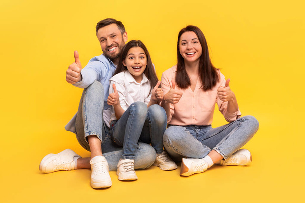 Happy family of three casually dressed giving thumbs up sitting together against yellow studio backdrop, parents and kid girl smiling to camera gesturing likes in approval of exciting offer - Photo, Image