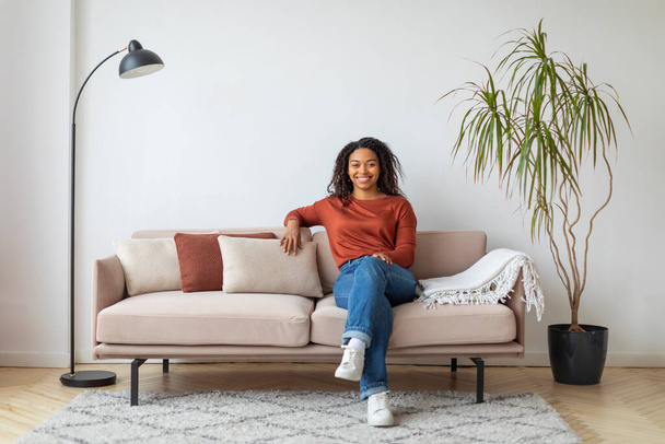 Веселая молодая черная женщина сидит комфортно на современном диване с подушками, счастливая африканская женщина тысячелетия позирует в хорошо освещенной гостиной, улыбается перед камерой, копирует пространство - Фото, изображение