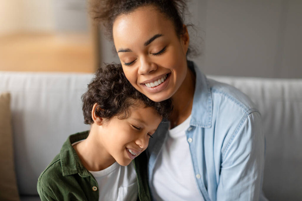 Radosny i czuły, czarny, kręcone włosy chłopiec otrzymuje ciepły uścisk od swojej promiennej matki, zarówno dzieląc szczerą chwilę miłości i bliskości na kanapie - Zdjęcie, obraz