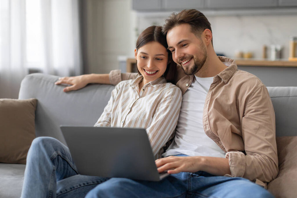 Feliz pareja joven cozied en el sofá, compartiendo momento alegre mientras miran la pantalla del ordenador portátil en el interior de la sala de estar bien iluminado en casa, navegar por Internet juntos - Foto, Imagen