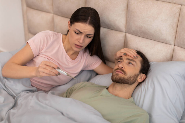 Znepokojená žena pozorně kontroluje horečku nemocného muže s teploměrem, ilustruje péči a starosti při nemoci v domácím prostředí - Fotografie, Obrázek