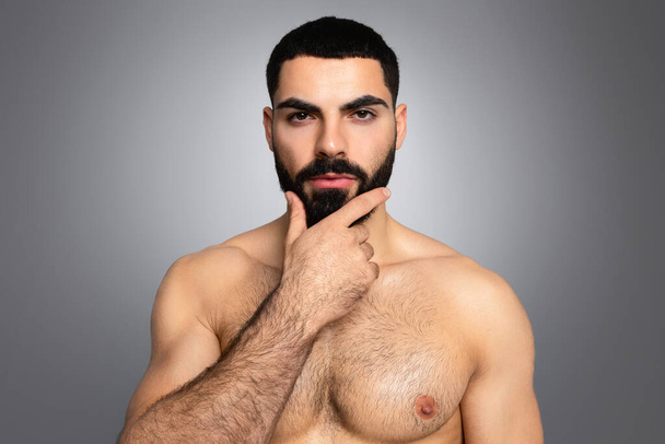 健康的なひげの概念のためのヒント. ハンサムなアスレチック裸ブルネット中東の若者は彼の厚いひげに触れ,灰色のスタジオの背景で孤立したカメラを見て,クローズアップ - 写真・画像