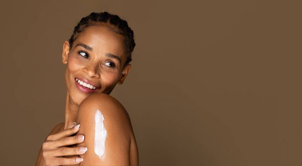 Promienna, afrykańska kobieta w średnim wieku, oglądająca się przez ramię z nałożeniem balsamu, ucieleśniająca pielęgnację skóry i pozytywność na brązowym tle, wolna przestrzeń - Zdjęcie, obraz