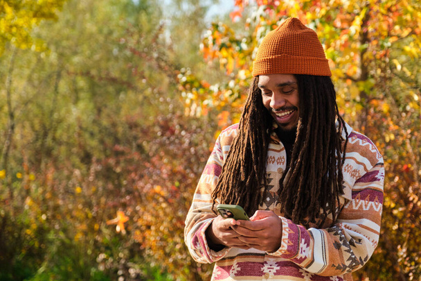 Afroamerykanin młody człowiek z dredami szczęśliwy korzystając ze smartfona w słoneczny dzień jesieni. - Zdjęcie, obraz