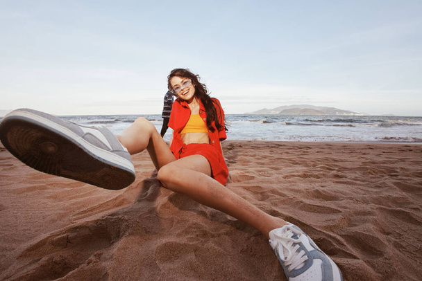 Happy Beach Fashion: uśmiechnięta kobieta w modnym stroju, siedzi na piasku z szerokim kątem widzenia na Morze letnie - Zdjęcie, obraz